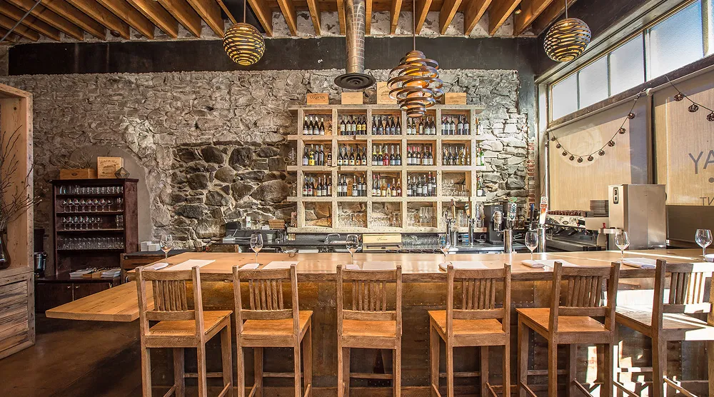 Cozy bar interior with wooden stools, a stone wall, shelves of liquor bottles, and hanging pendant lights.