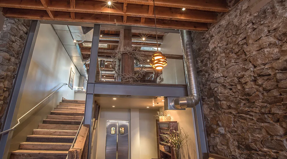 Interior staircase with wooden steps, rustic stone wall, exposed wooden ceiling beams, and industrial metal ductwork.
