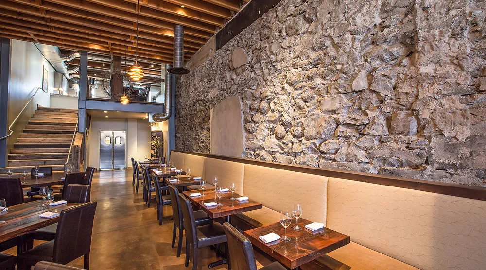 Interior of a restaurant with wooden tables, leather chairs, beige bench seating along a stone wall, and exposed wooden ceiling beams.