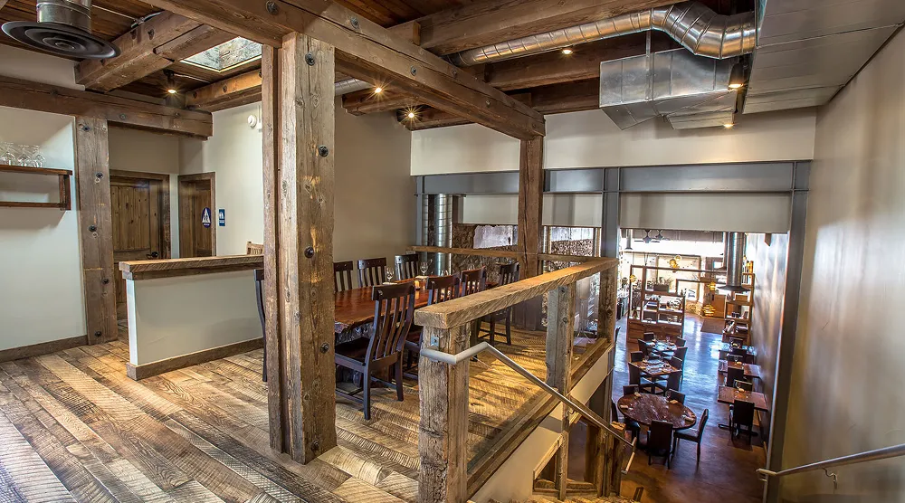 Interior of a rustic restaurant with wooden beams, tables, and chairs, featuring a staircase leading down to more seating.