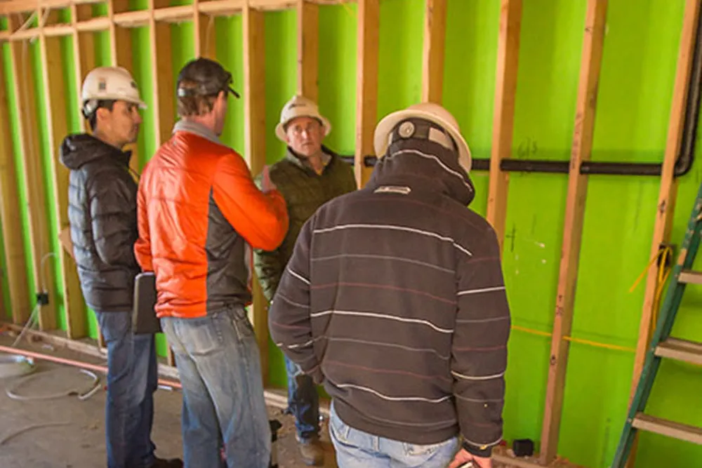 Four construction workers wearing hard hats discussing plans inside a building under construction with exposed wooden studs and green insulation walls.