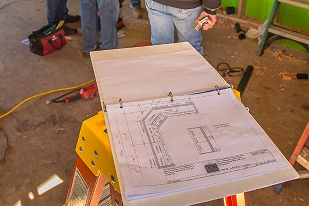 Open binder with construction plans on a yellow stepladder in a workshop with people standing nearby.