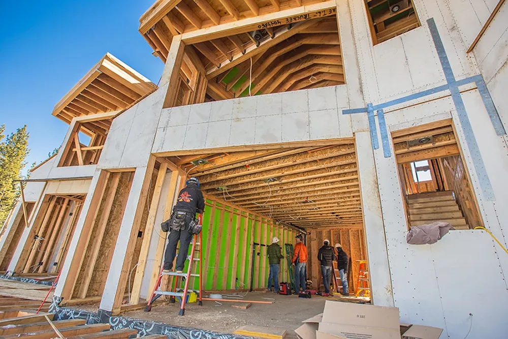 Construction workers working on framing the interior of a large wooden house under clear blue sky.
