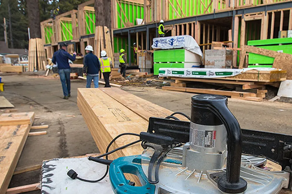 Construction site with stacked wooden boards and power tool in foreground and workers inspecting a partially framed building in the background.