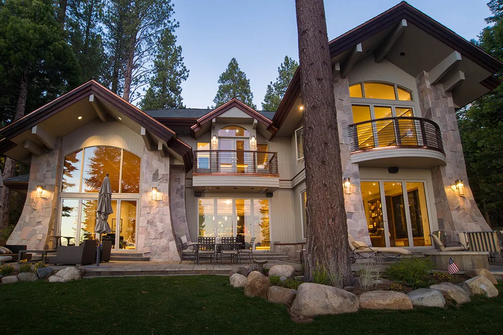 Modern two-story house with large windows and balconies, surrounded by tall pine trees at dusk.