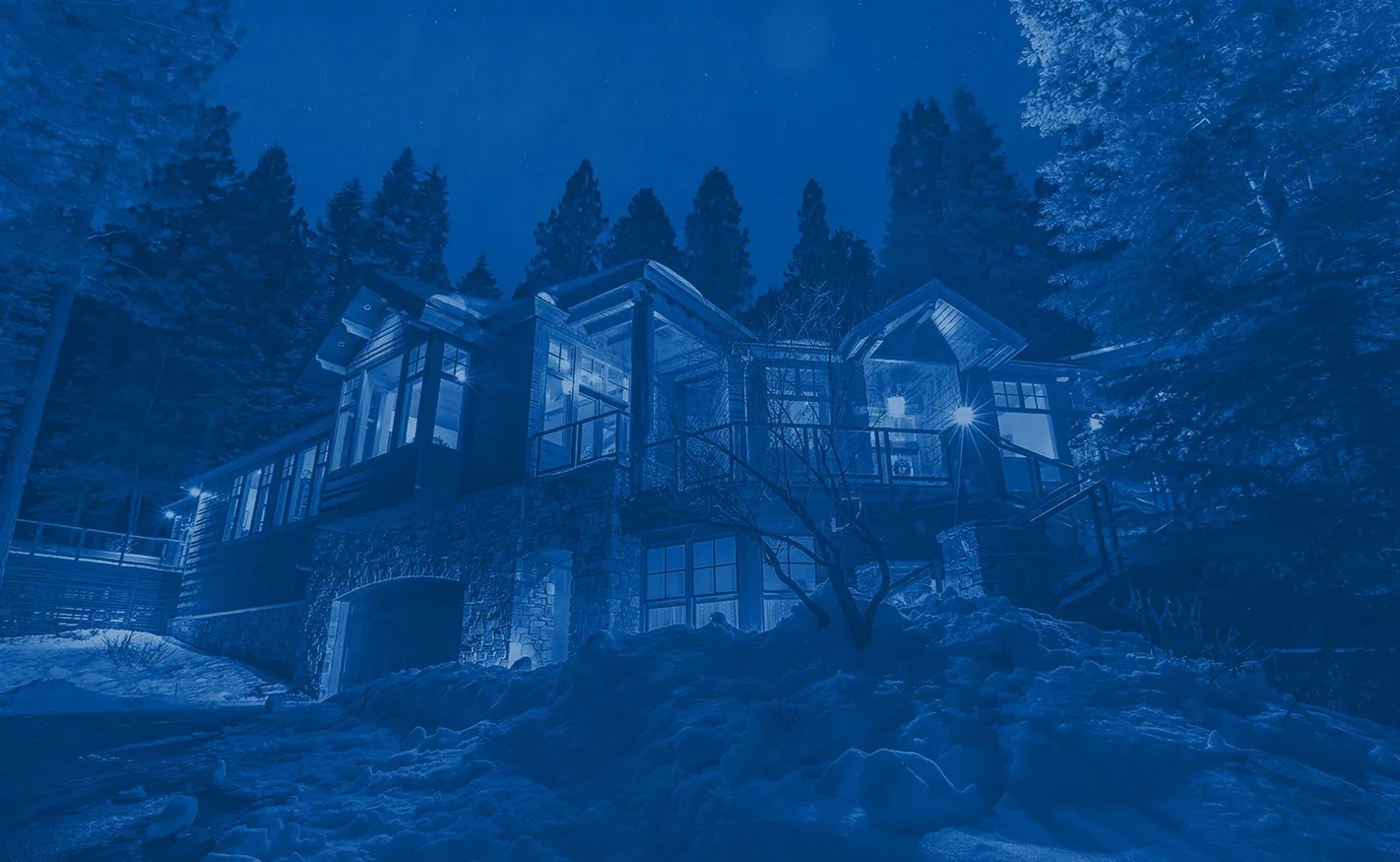 Stone and wood cabin illuminated at night surrounded by snow and tall pine trees.
