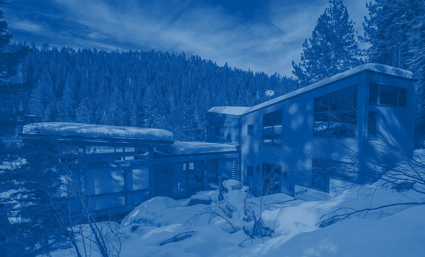 Modern house covered with snow in a forested mountainous area under a partly cloudy sky.