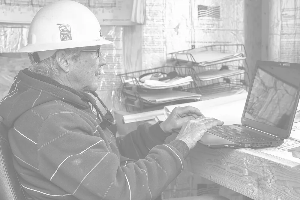 Construction worker wearing a hard hat working on a laptop at a desk with stacked papers and an American flag in the background.
