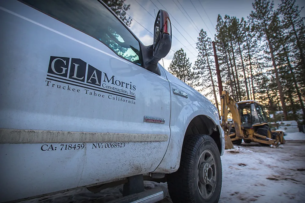 Side view of a white pickup truck with GLA Morris Construction, Inc. logo and license numbers parked on snowy ground near a yellow backhoe and pine trees at sunset.