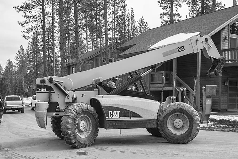 CAT TH460B telescopic handler parked on a road in front of a wooden cabin with trees in the background.