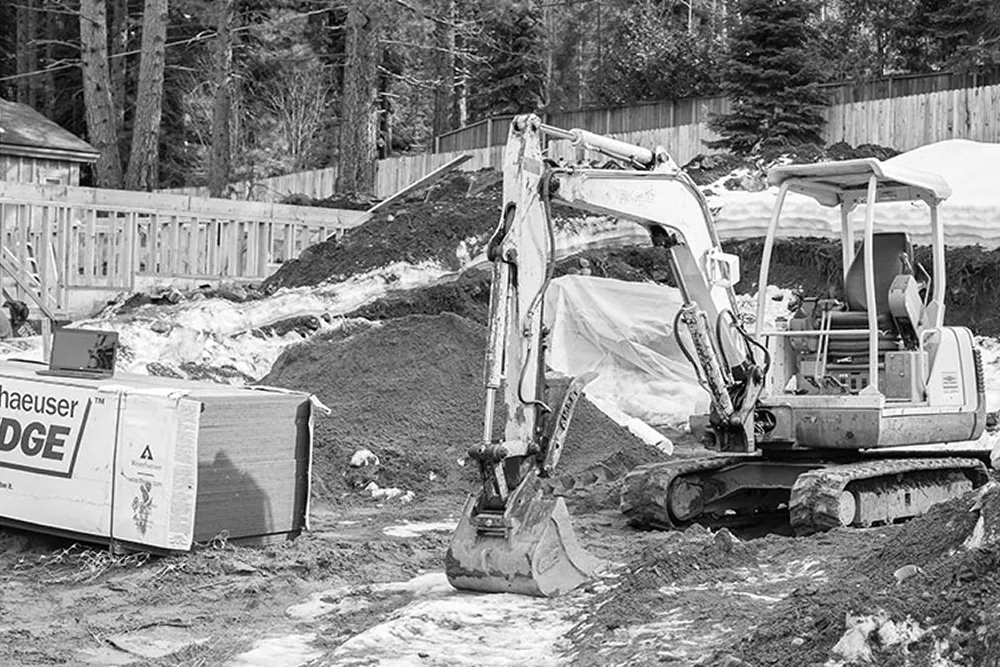 Mini excavator parked on a snowy and muddy construction site with piles of dirt and large construction materials nearby.