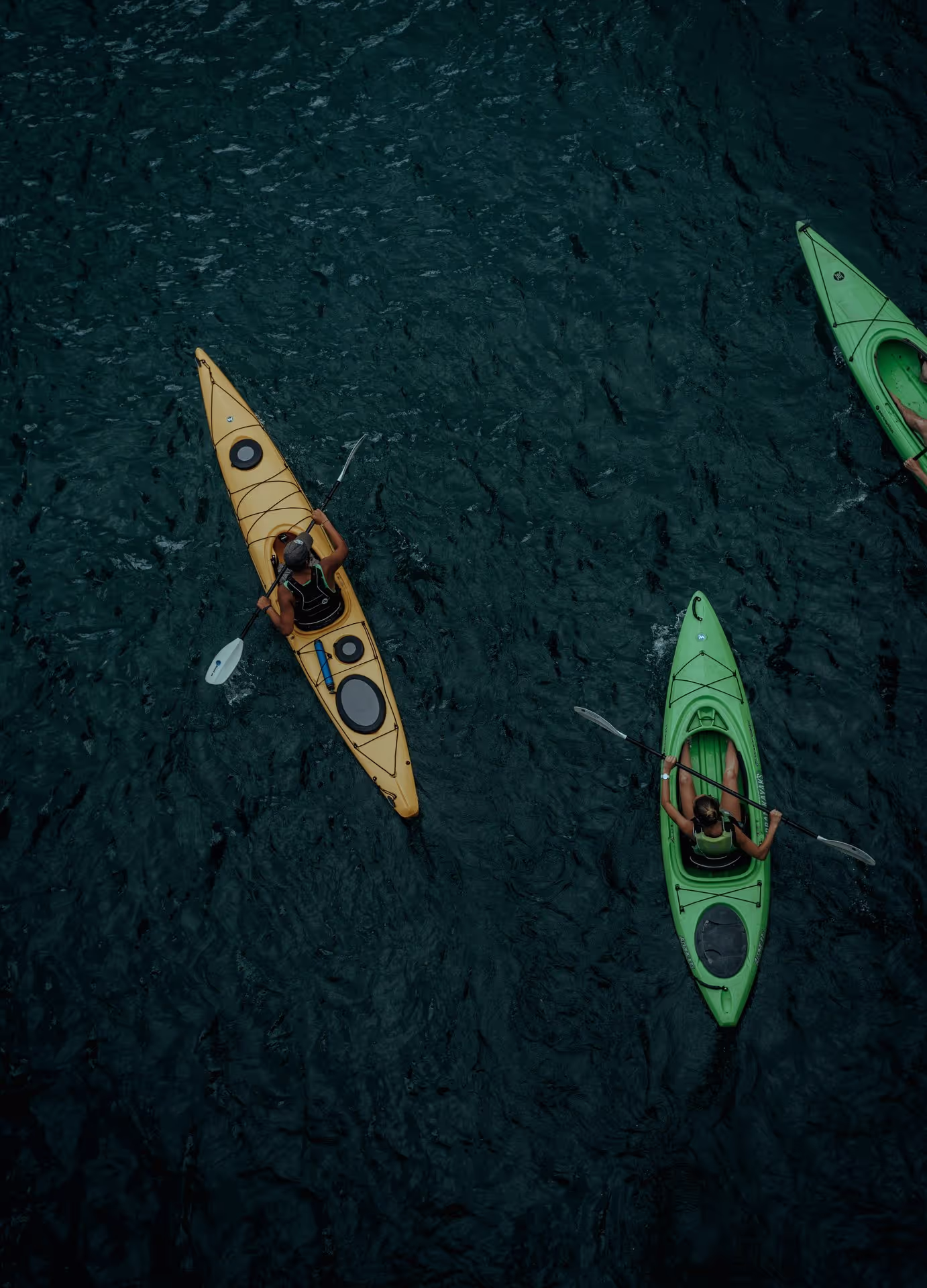Aerial view of three kayakers in green and yellow kayaks paddling on dark blue water, creating gentle ripples around them.