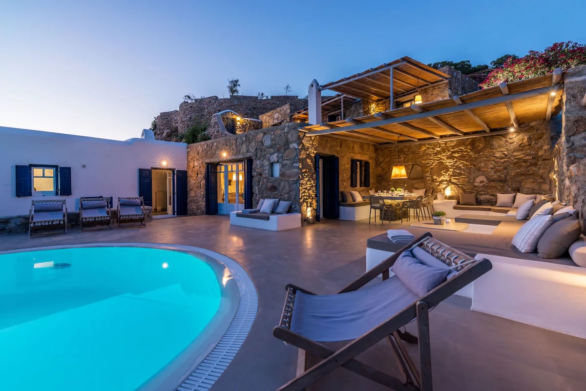 Cozy Mykonos villa at dusk with a lit pool, stone walls, and outdoor seating area.