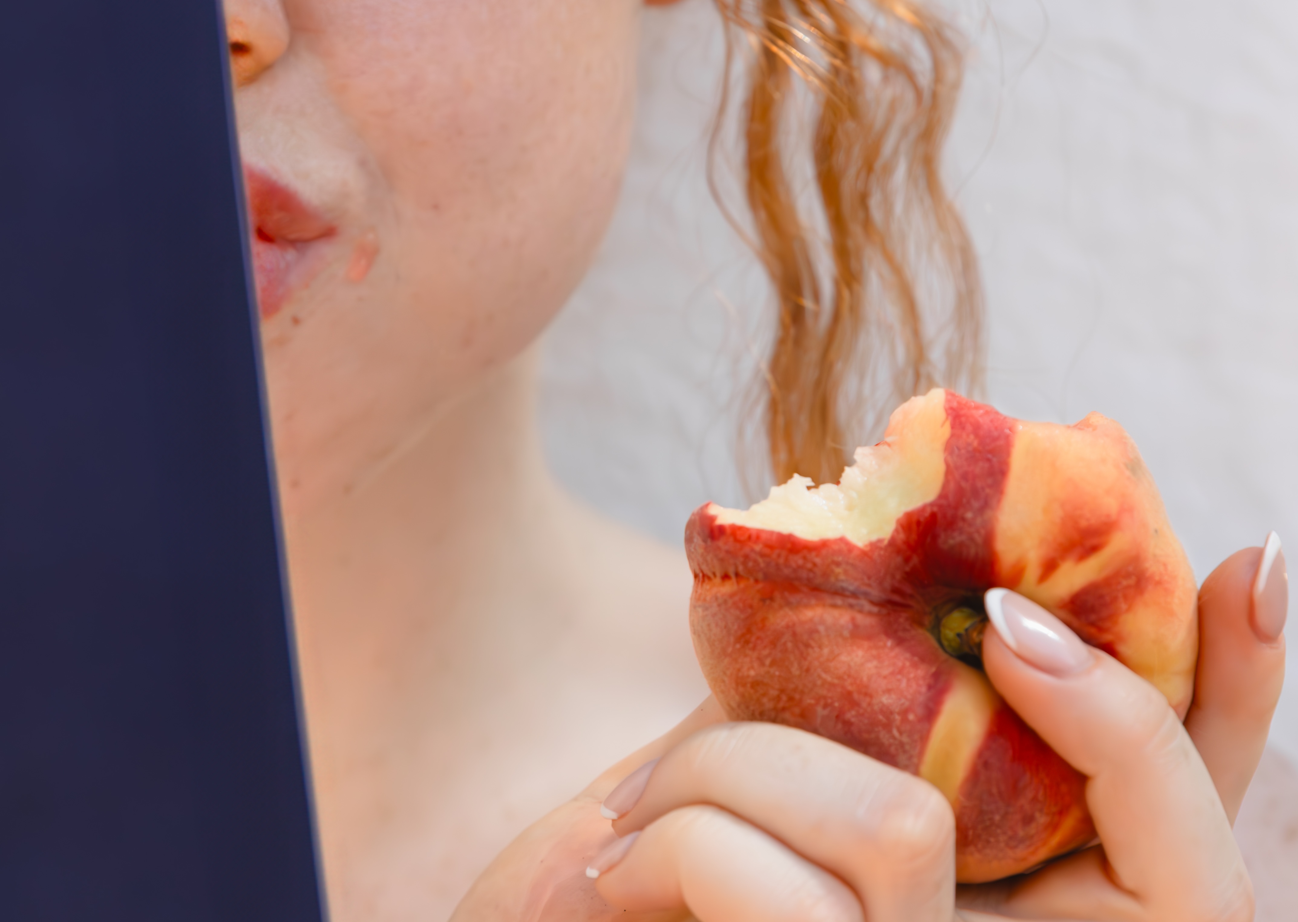 A close-up of a woman with long red hair holding a peach with a bite taken out of it.