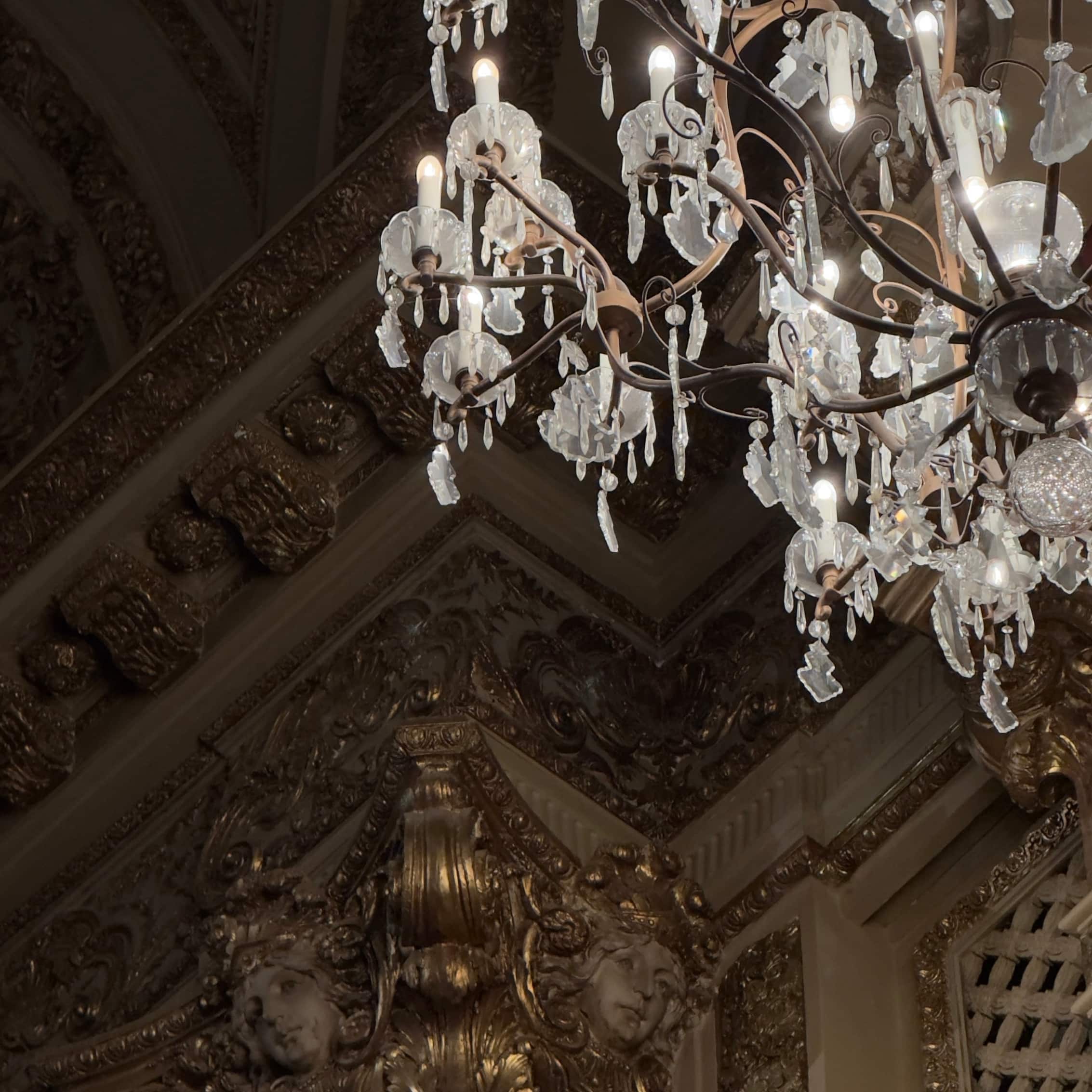 A crystal chandelier in front of a lavish and ornate gold wall decoration.
