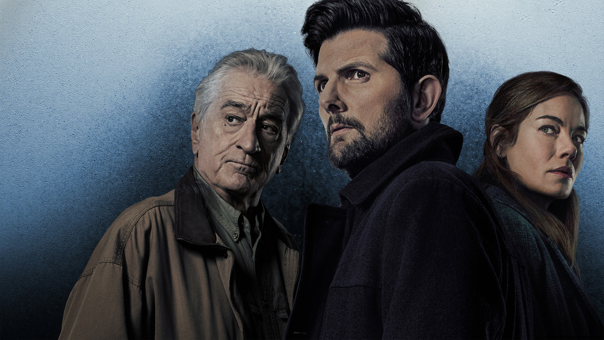 Three serious-looking people in dark clothing stand against a textured blue background: an older grey-haired man on the left, a dark-haired bearded man in the centre, and a long-haired woman on the right.