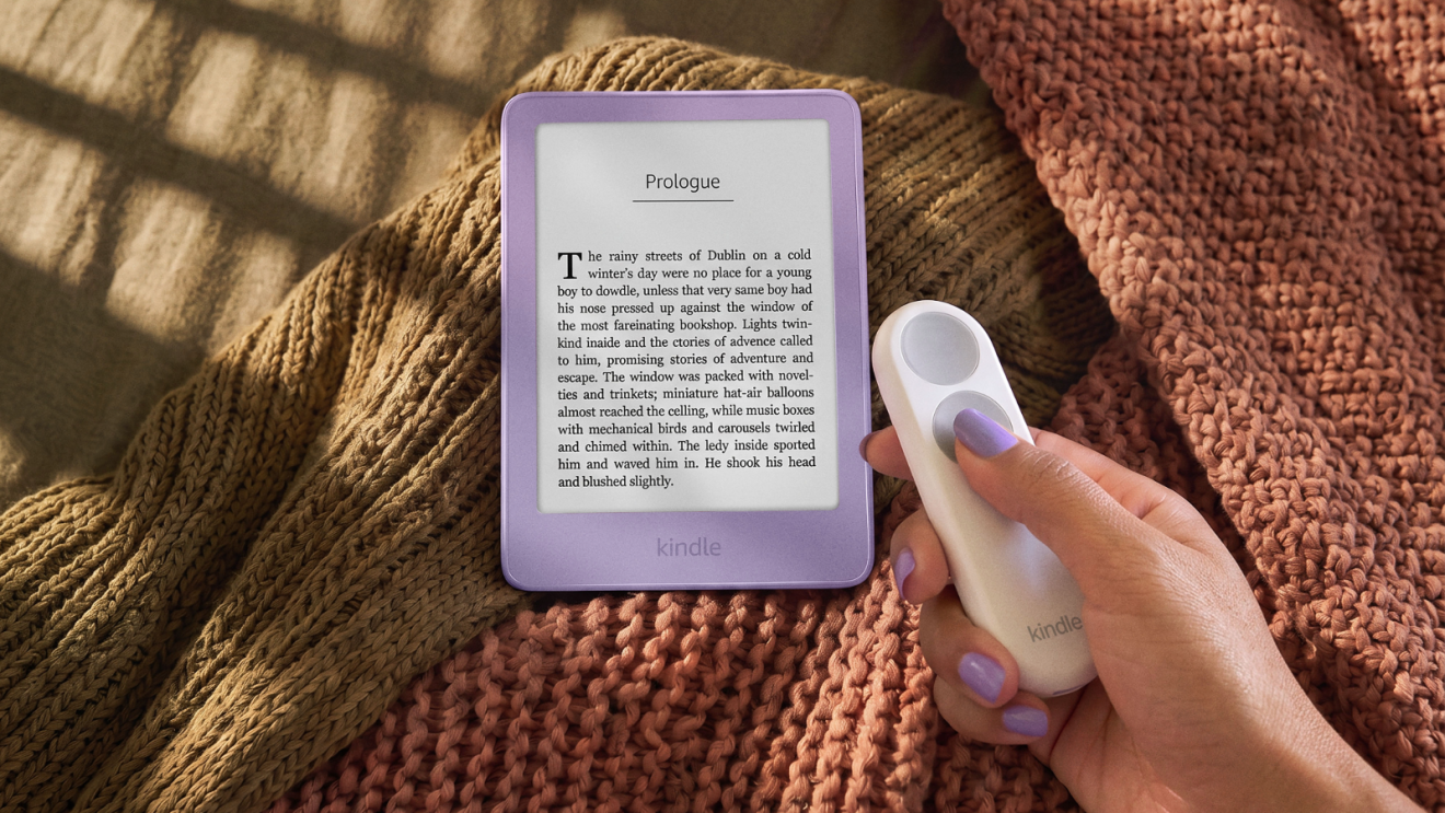 A light purple Amazon Kindle resting on a blanket with a hand in front of it, holding a remote clicker to turn the pages.