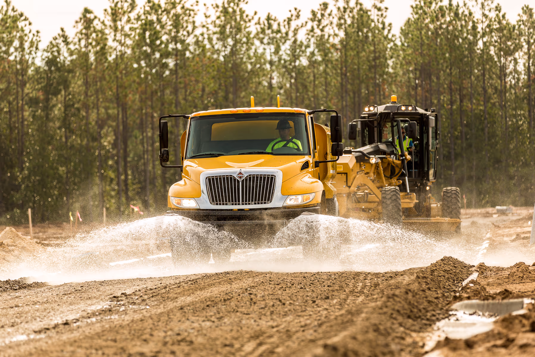 Yellow truck and grader kicking up dust on dirt construction site