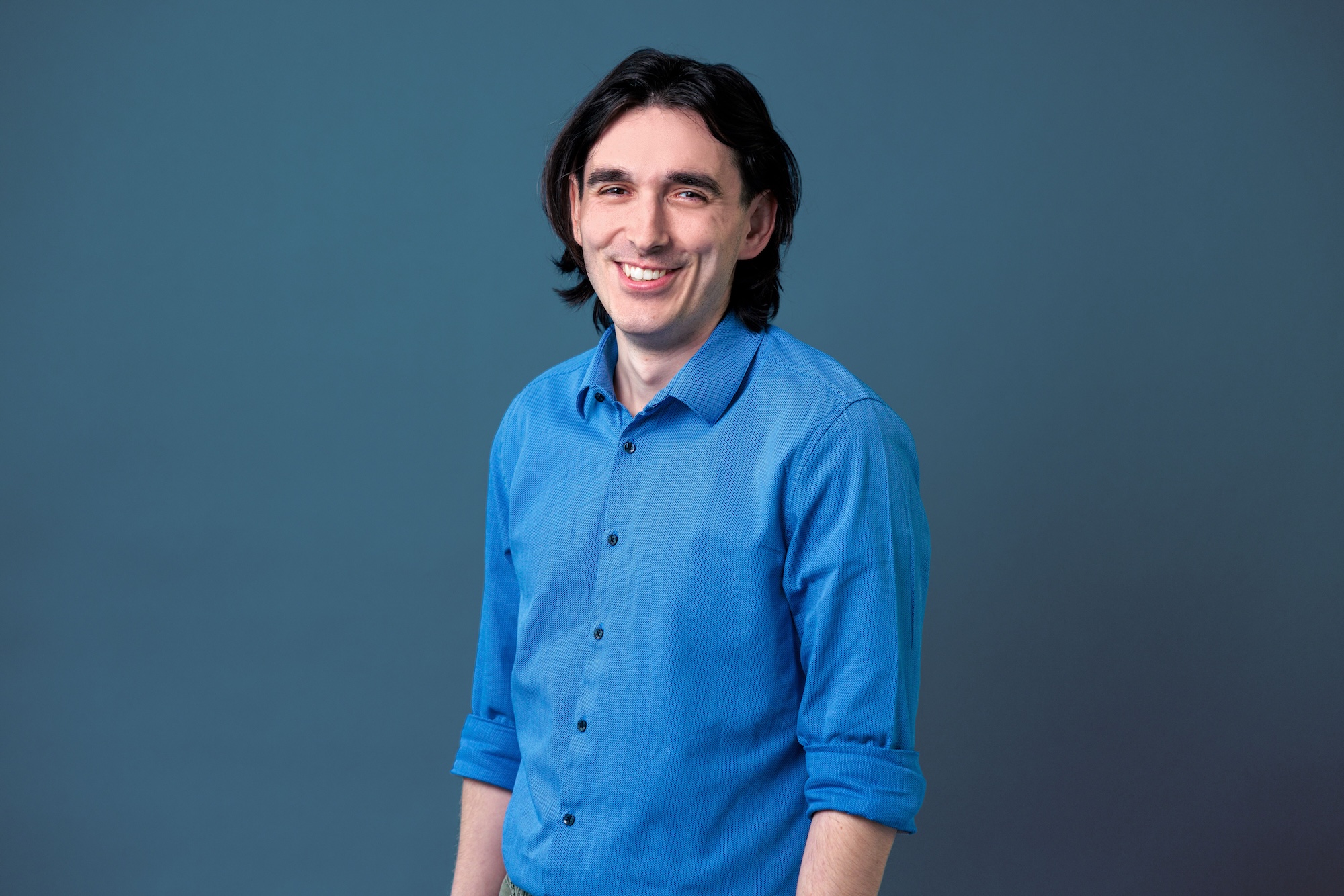 Junger Mann mit dunklen Haaren in blauem Hemd steht vor einfarbigem graublauem Hintergrund und lächelt.