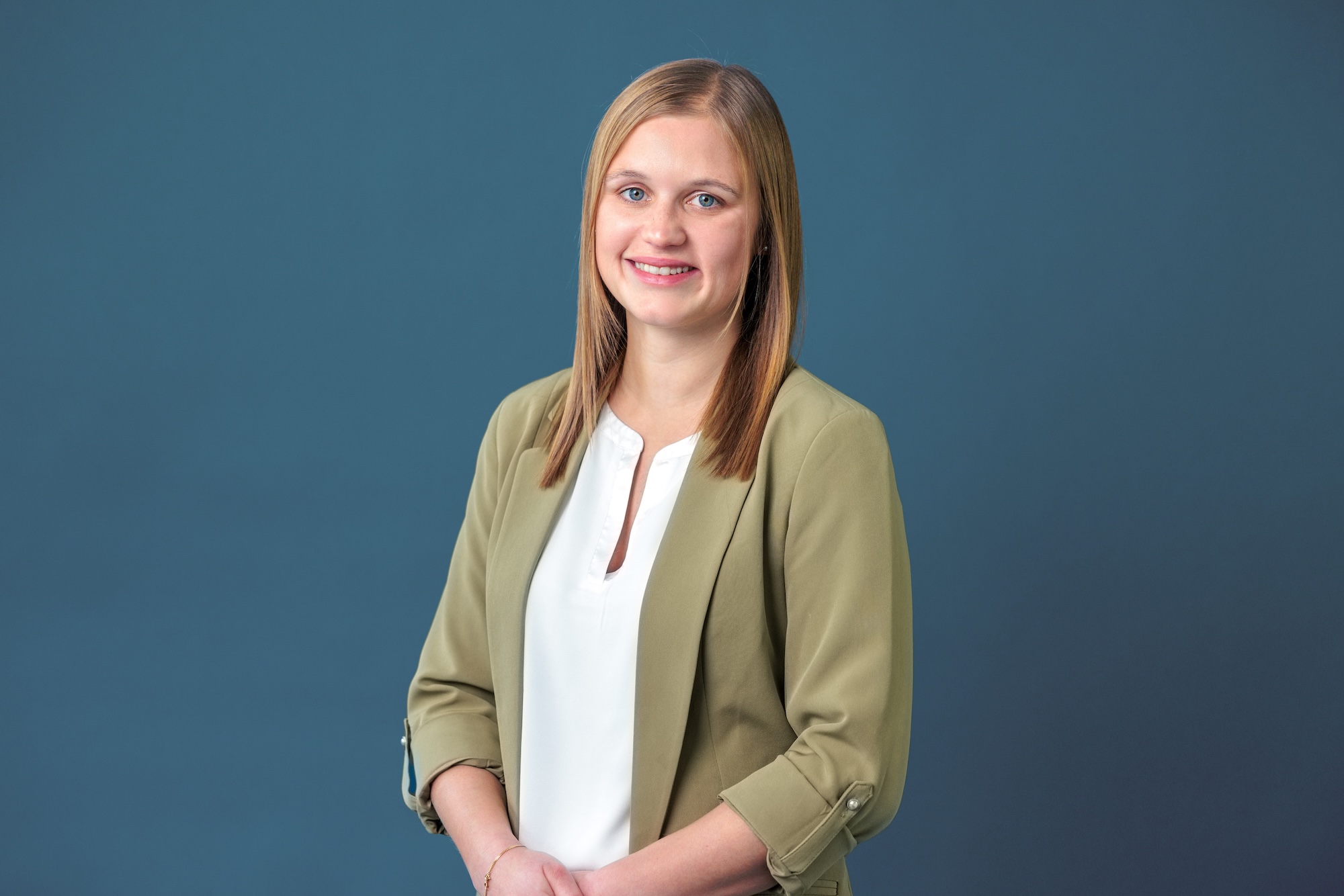 Porträt einer lächelnden jungen Frau mit langen blonden Haaren, die eine olivgrüne Jacke und eine weiße Bluse trägt, vor blauem Hintergrund.