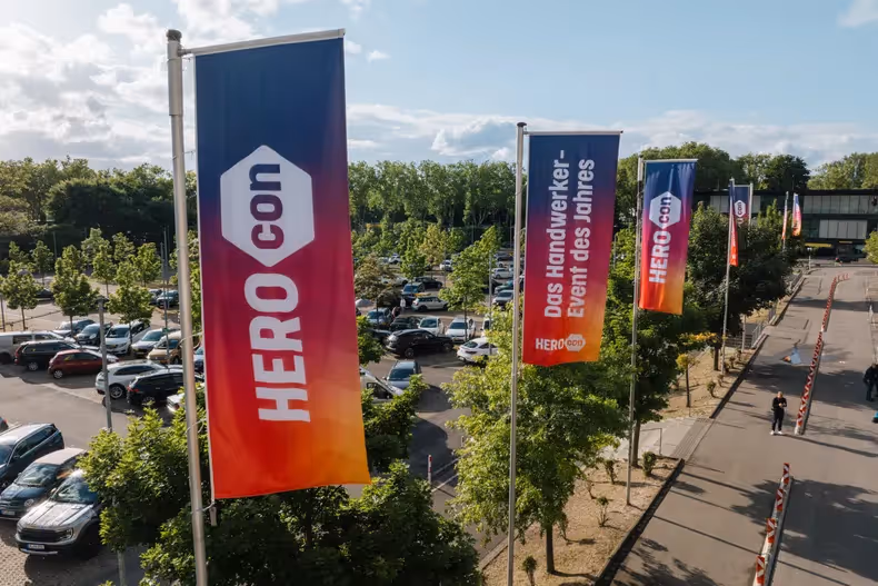 HEROcon-Fahnen mit Farbverlauf an Straße neben Parkplatz; eine mit „Das Handwerker-Event des Jahres“ unter hellem, teils bewölktem Himmel.