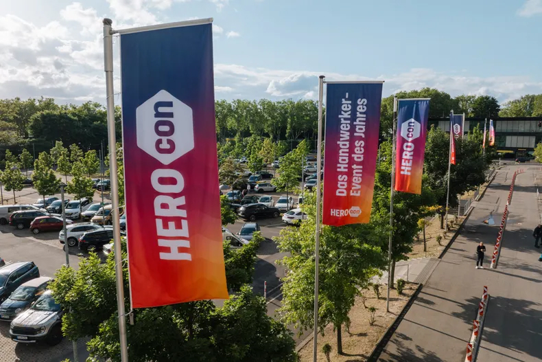 HEROcon-Fahnen mit Farbverlauf an Straße neben Parkplatz; eine mit „Das Handwerker-Event des Jahres“ unter hellem, teils bewölktem Himmel.