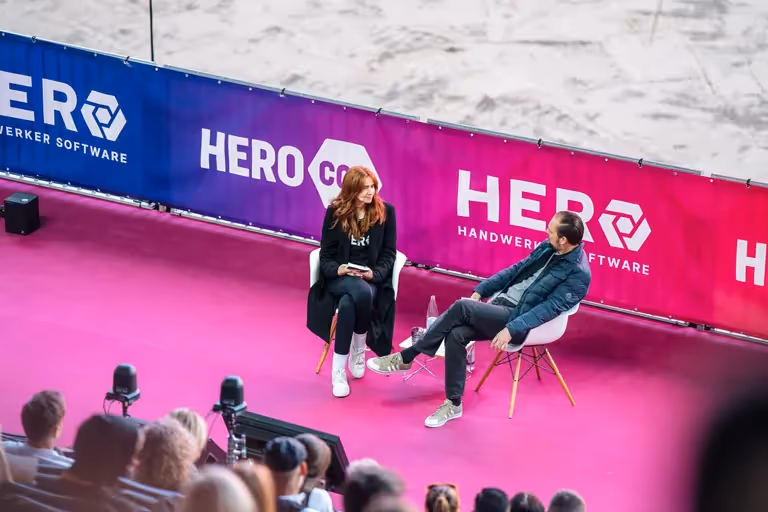 Zwei Personen im Gespräch auf Bühne mit HEROcon-Banner; Publikum im Vordergrund, pinker Teppich.