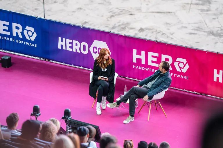 Zwei Personen im Gespräch auf Bühne mit HEROcon-Banner; Publikum im Vordergrund, pinker Teppich.