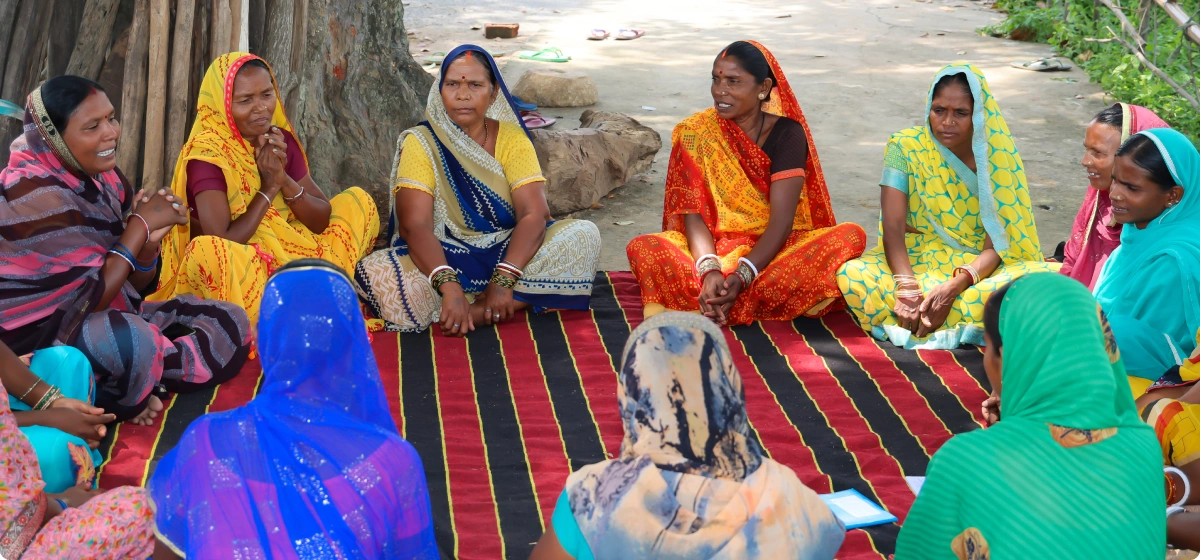 A group of women dressed in colorful traditional Indian sarees sitting in a circle on a striped red and black mat outdoors.