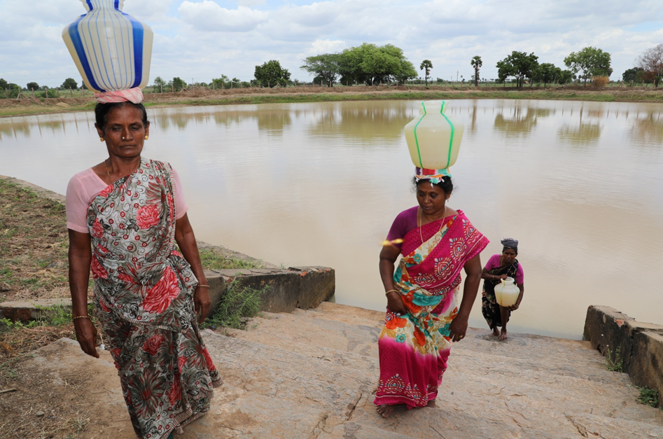 Three women dressed in colorful saris carrying large water containers on their heads walking up concrete steps from a pond under a cloudy sky.