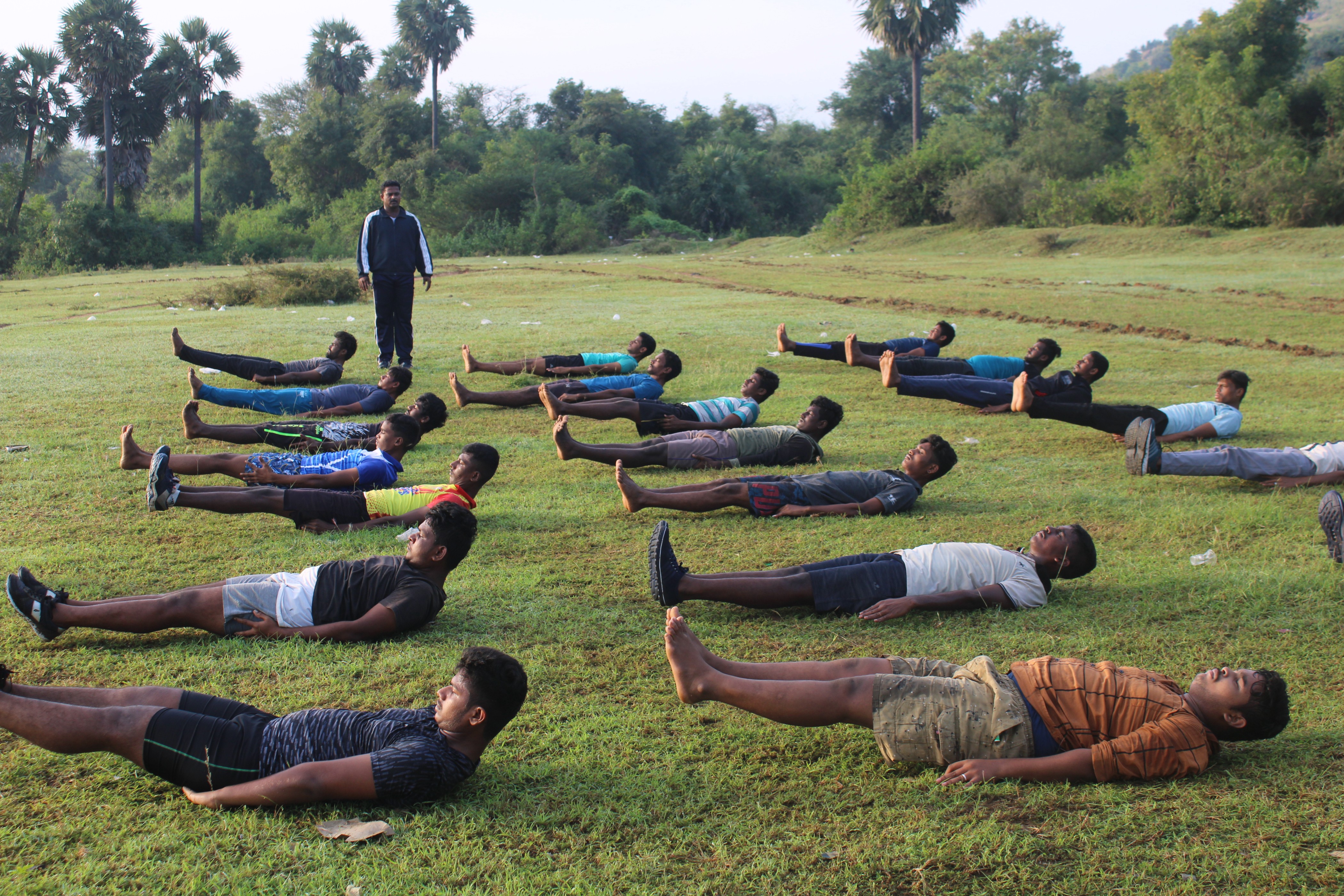 Group of young men lying on grass outdoors performing leg-raise exercises with an instructor standing nearby.