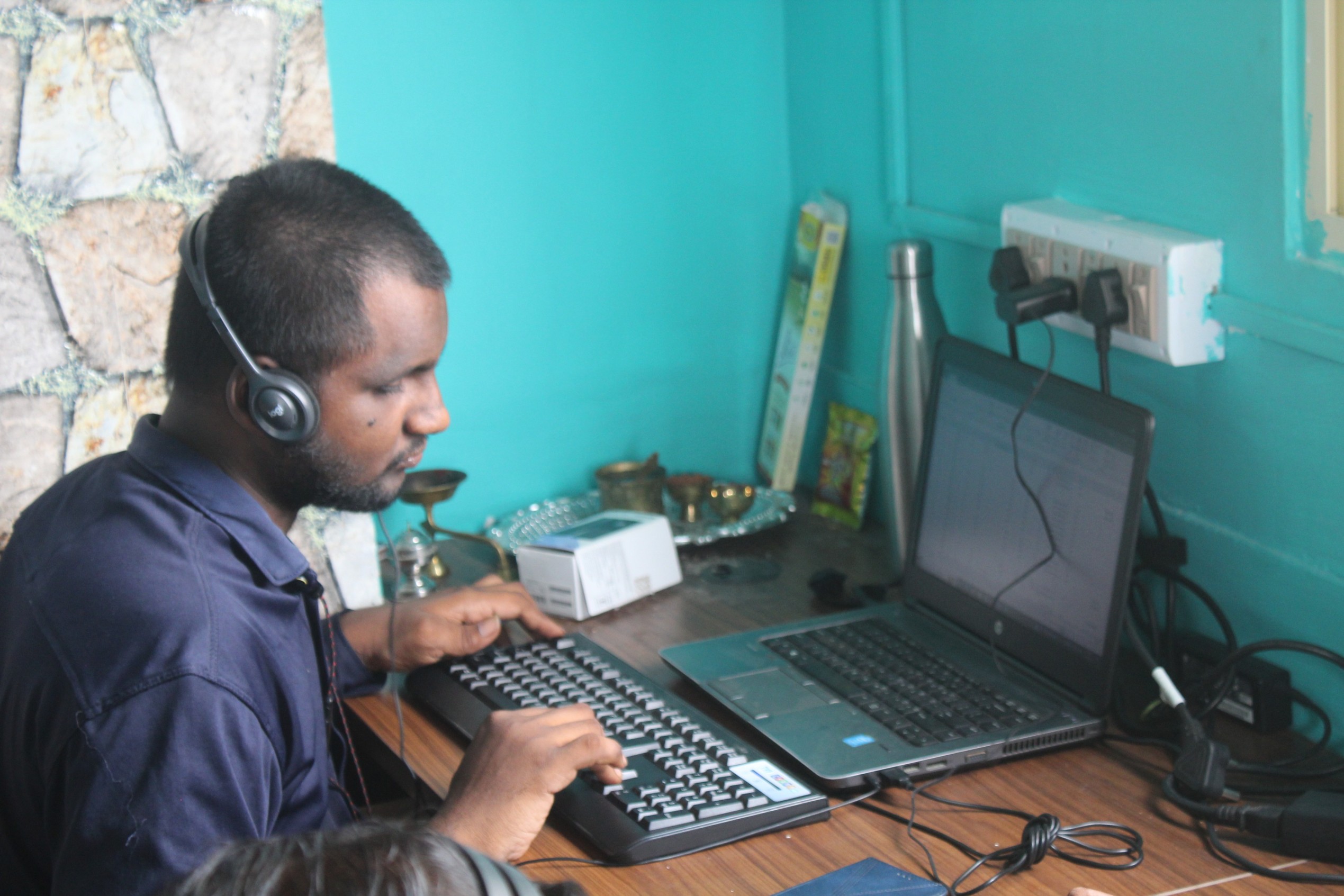 Man wearing headphones typing on a wireless keyboard connected to a laptop on a wooden desk in a turquoise room.