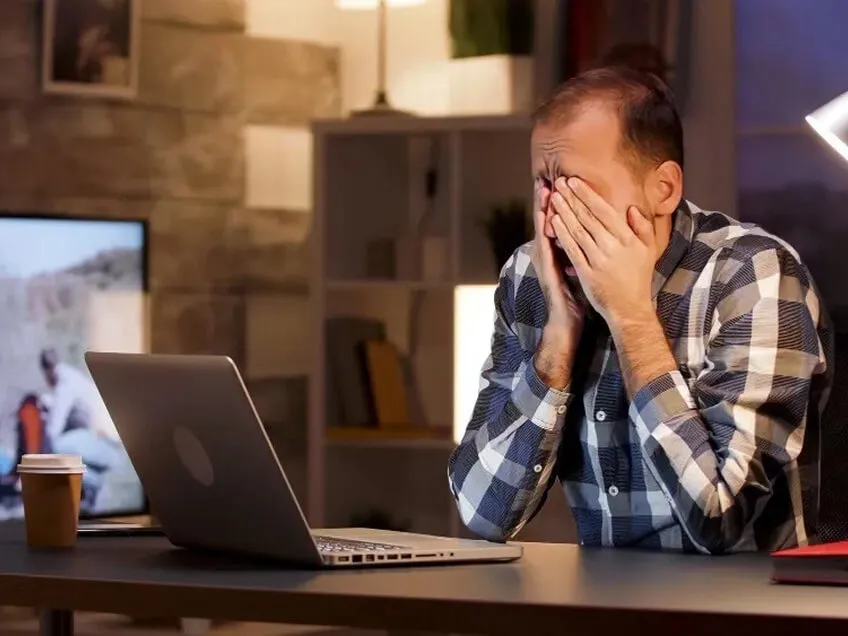 Man at a desk covering his face with both hands in frustration.