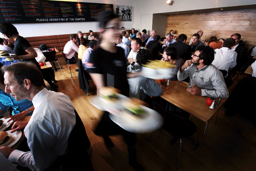 A blurred server carrying plates through a busy restaurant with many seated customers dining.