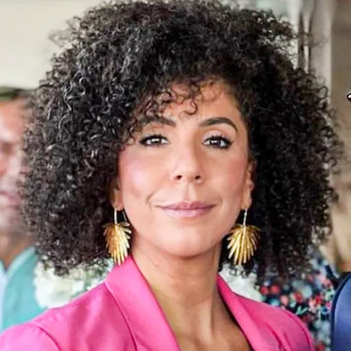 a woman with curly hair wearing a pink jacket and large gold earrings