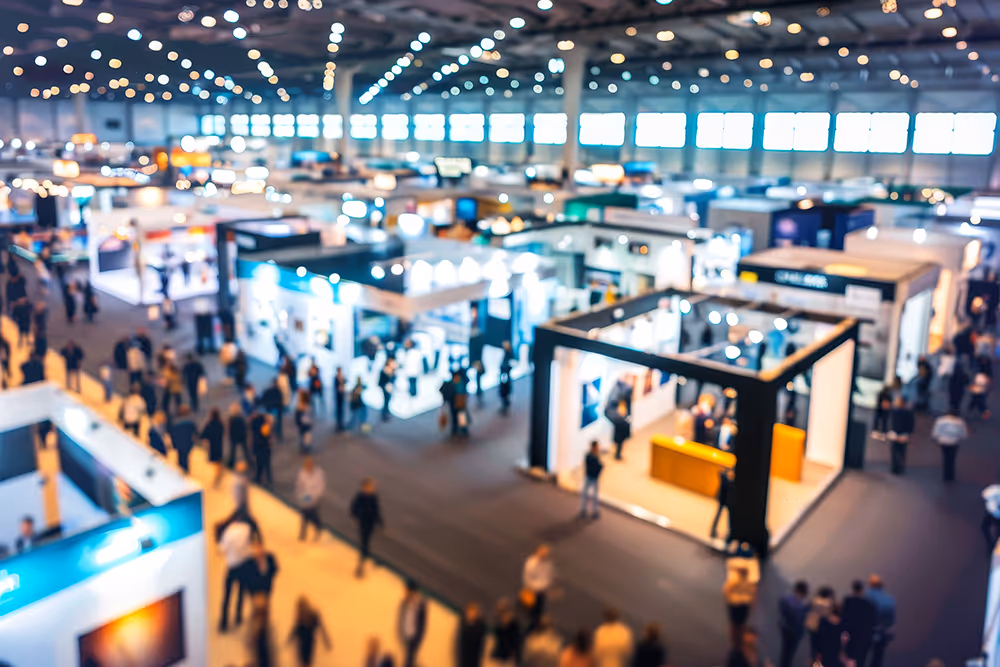 Blurred wide view of a busy exhibition hall with booths and people walking around.