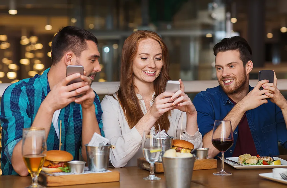 Three friends sitting at a restaurant table, smiling and using smartphones with burgers and drinks in front of them.