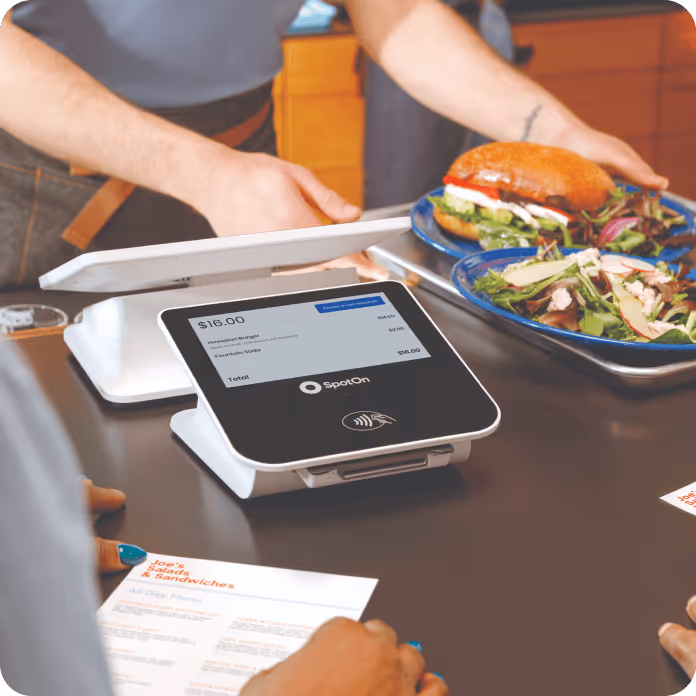 Person placing two plates with a burger and salad on a counter next to a SpotOn touchscreen payment terminal displaying a $16.00 total.