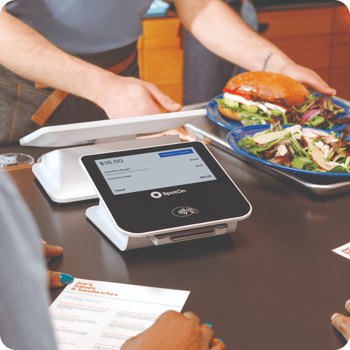 Person placing two plates with a burger and salad on a counter next to a SpotOn touchscreen payment terminal displaying a $16.00 total.