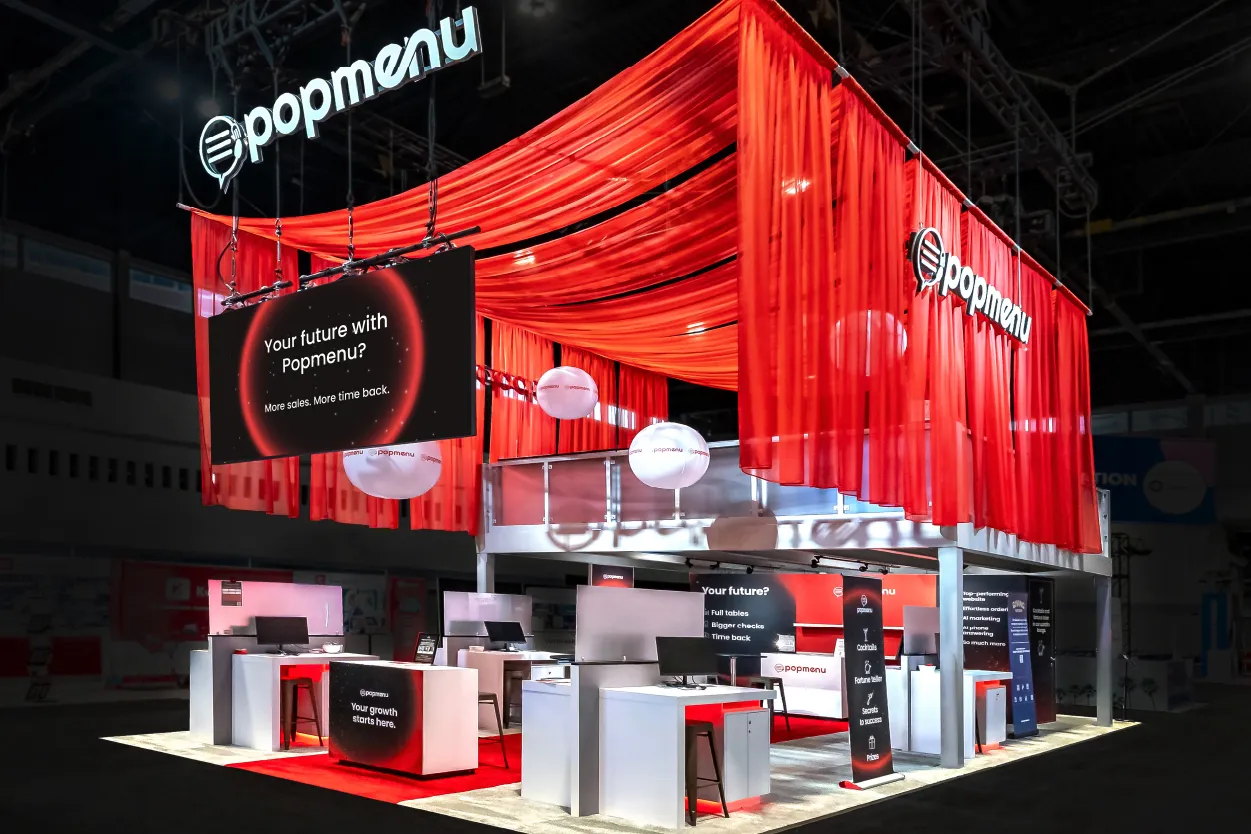 Popmenu trade show booth with red draped fabric, hanging signage, white desks, and digital screens promoting sales growth and time savings.