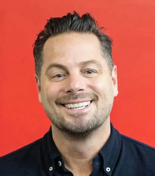Smiling man with short dark hair and trimmed beard wearing a dark shirt against a red background.