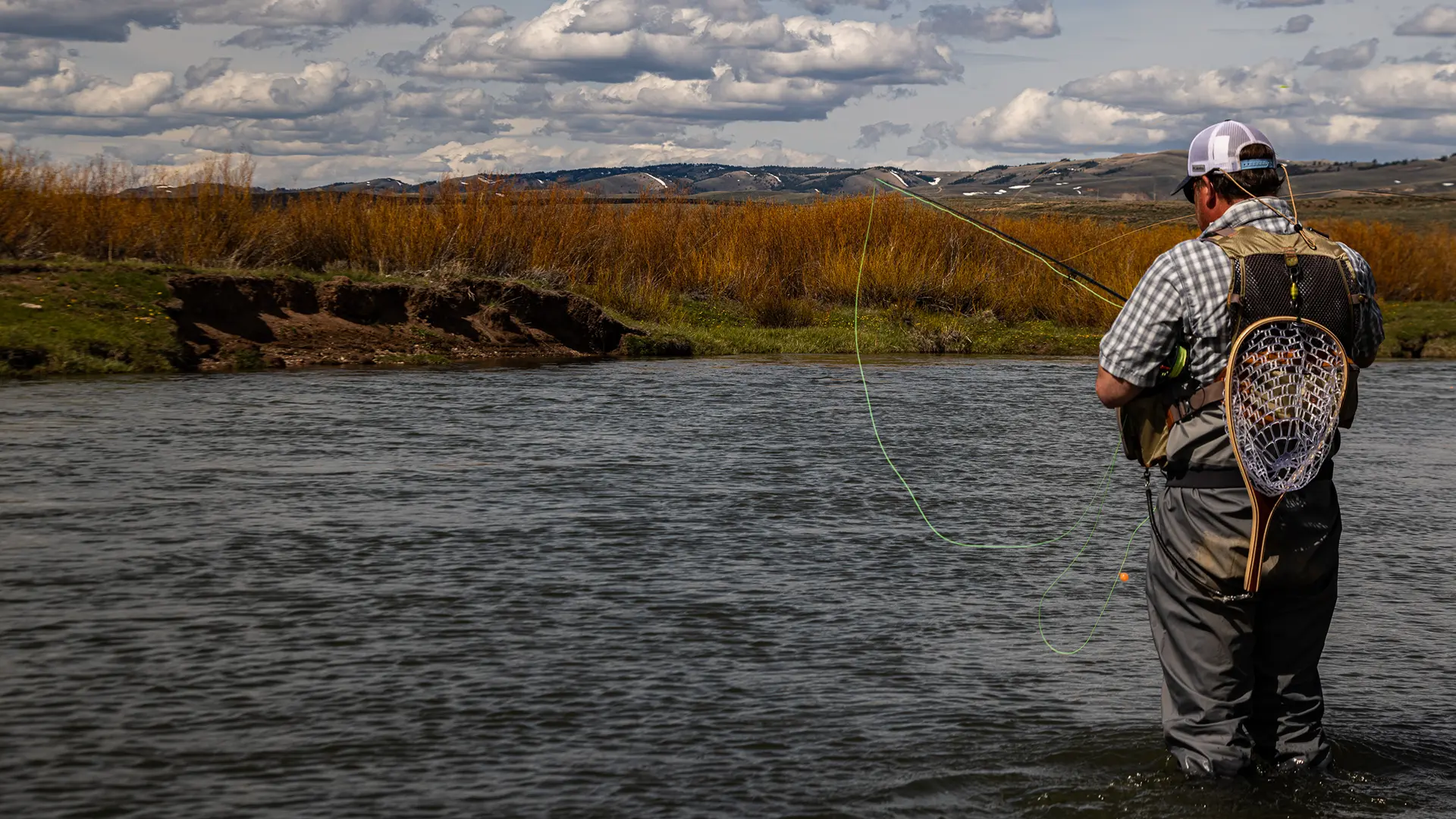 Man fly fishing in a river with mountains and rolling hills in the distance, under a partly cloudy sky in Heber Valley, Utah.