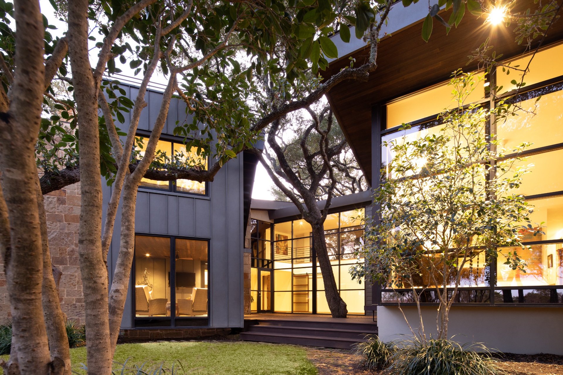 Modern custom home exterior with large glass windows, natural stone walls, and surrounding oak trees at dusk – River City Construction and Design project in Austin, Texas.