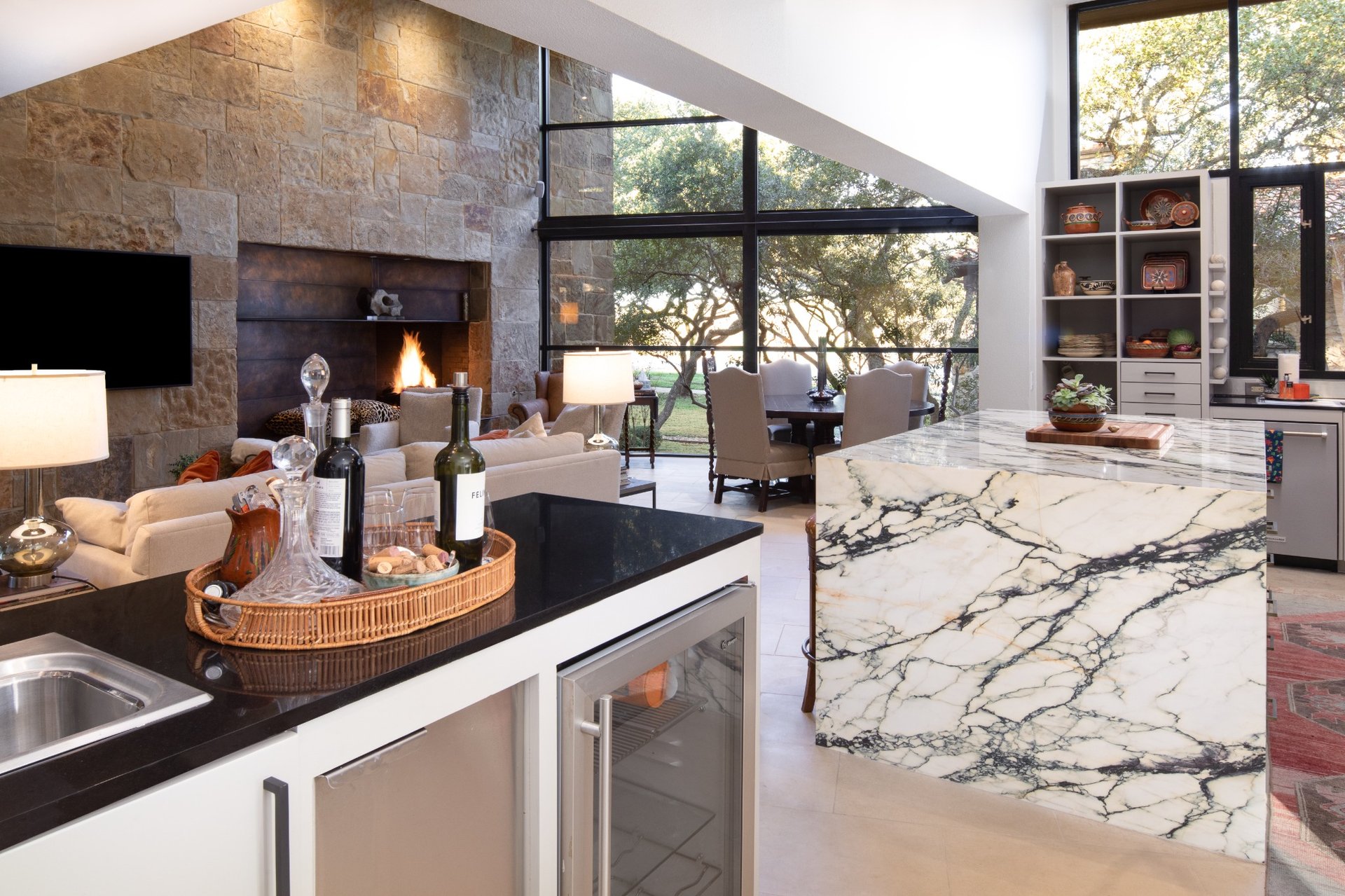 Open-concept living area with stone fireplace, marble kitchen island, and floor-to-ceiling windows overlooking Texas oak trees – River City Construction and Design.