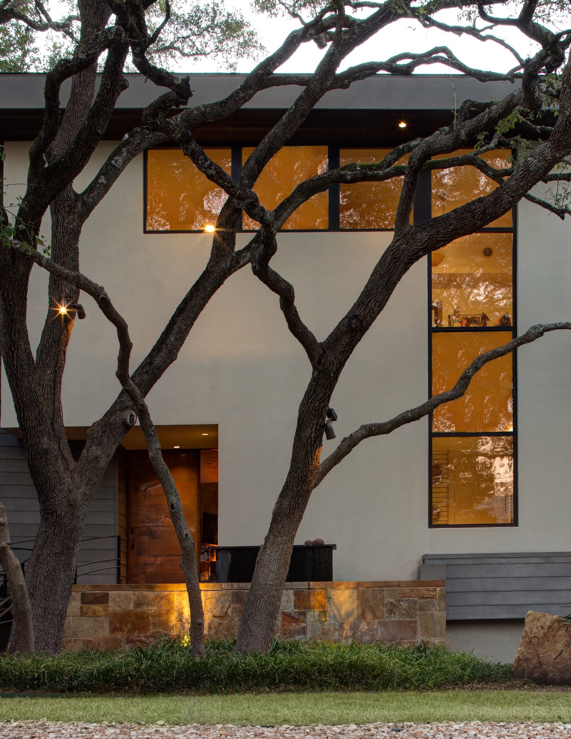  Modern Texas home front exterior with large oak trees, custom steel door, and warm architectural lighting – River City Construction and Design.