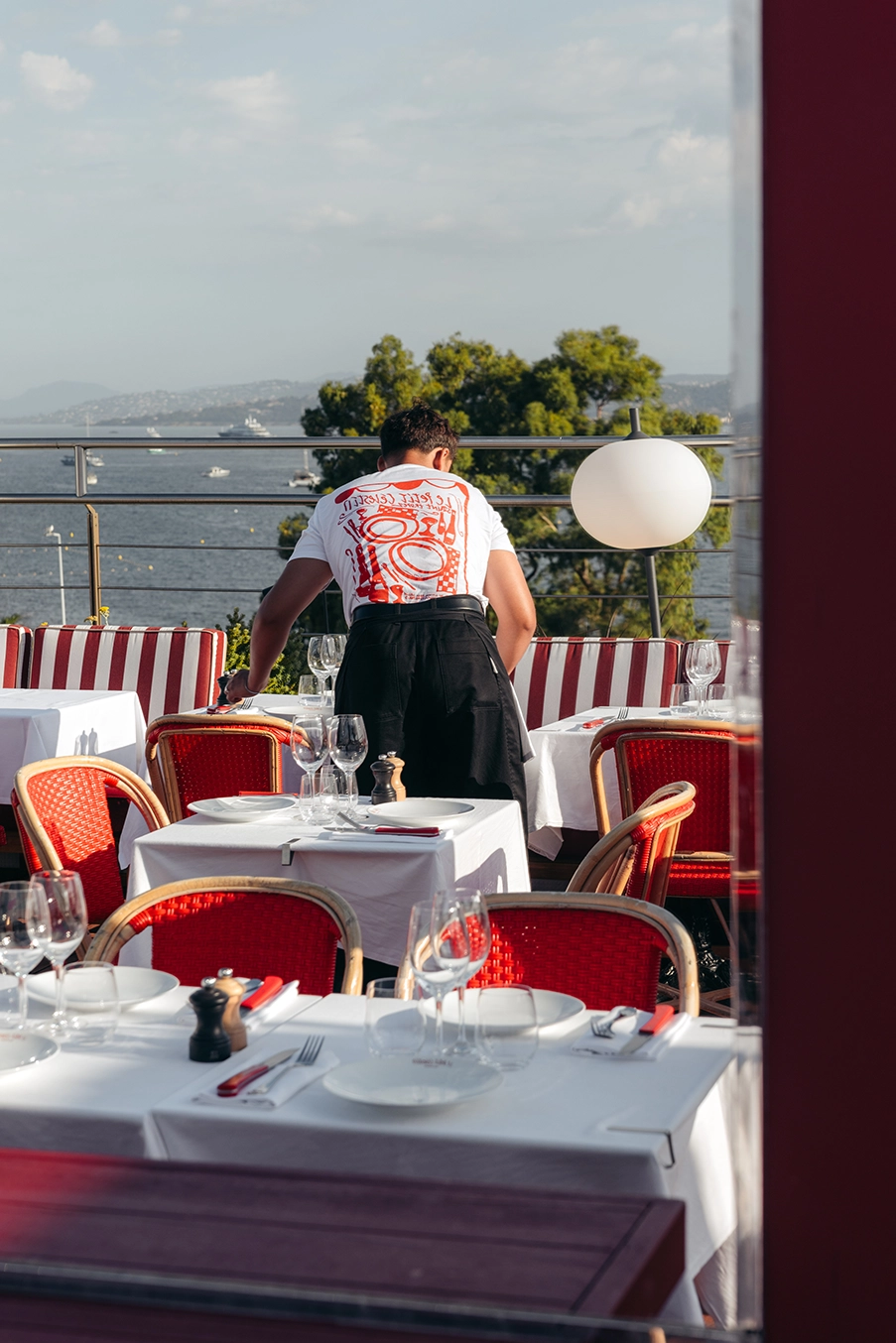 Serveur prépare une table en terrasse avec chaises rouges, vue sur la mer et le ciel clair.