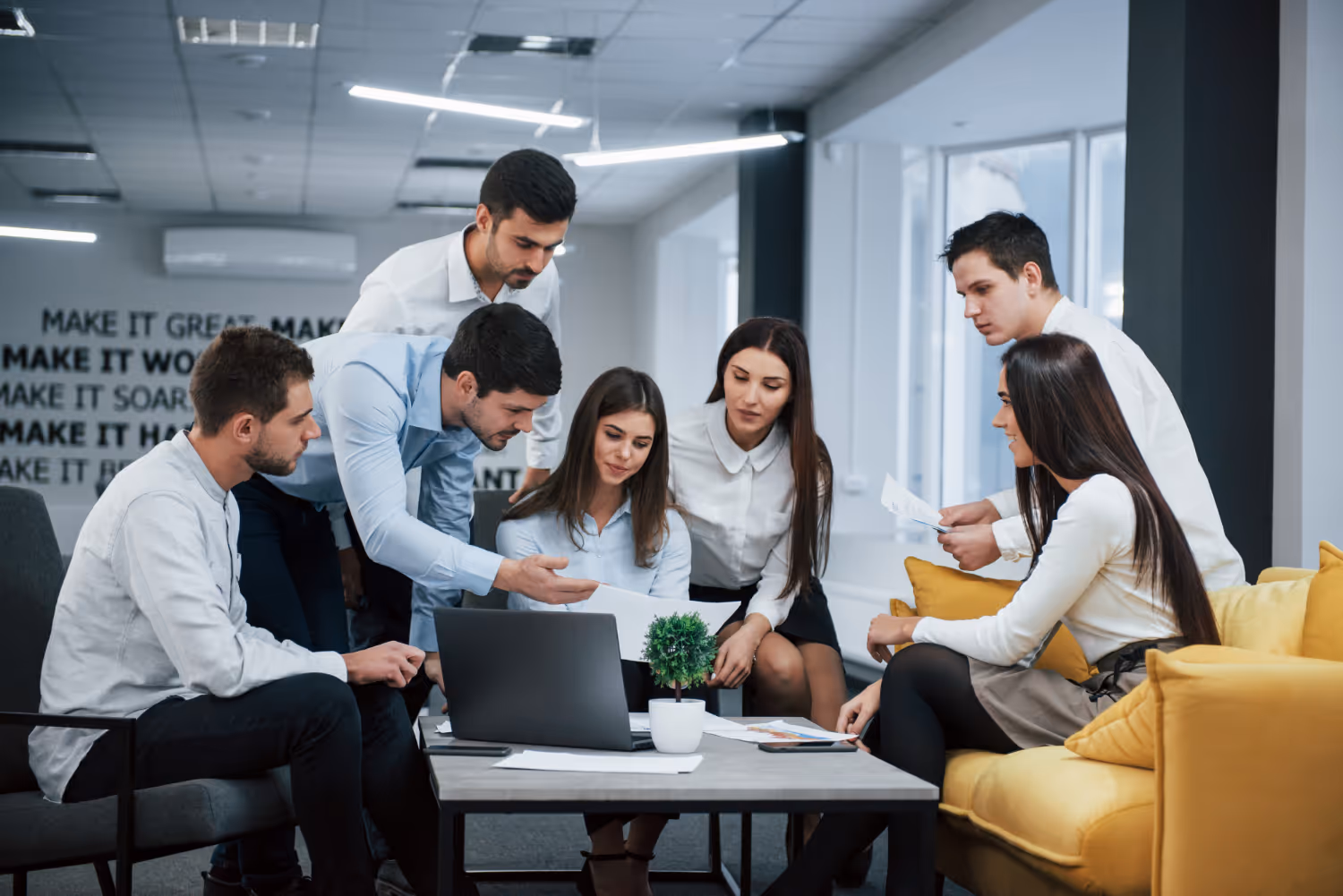 A group of seven people are collaborating around a table in an office, looking at papers and a laptop.