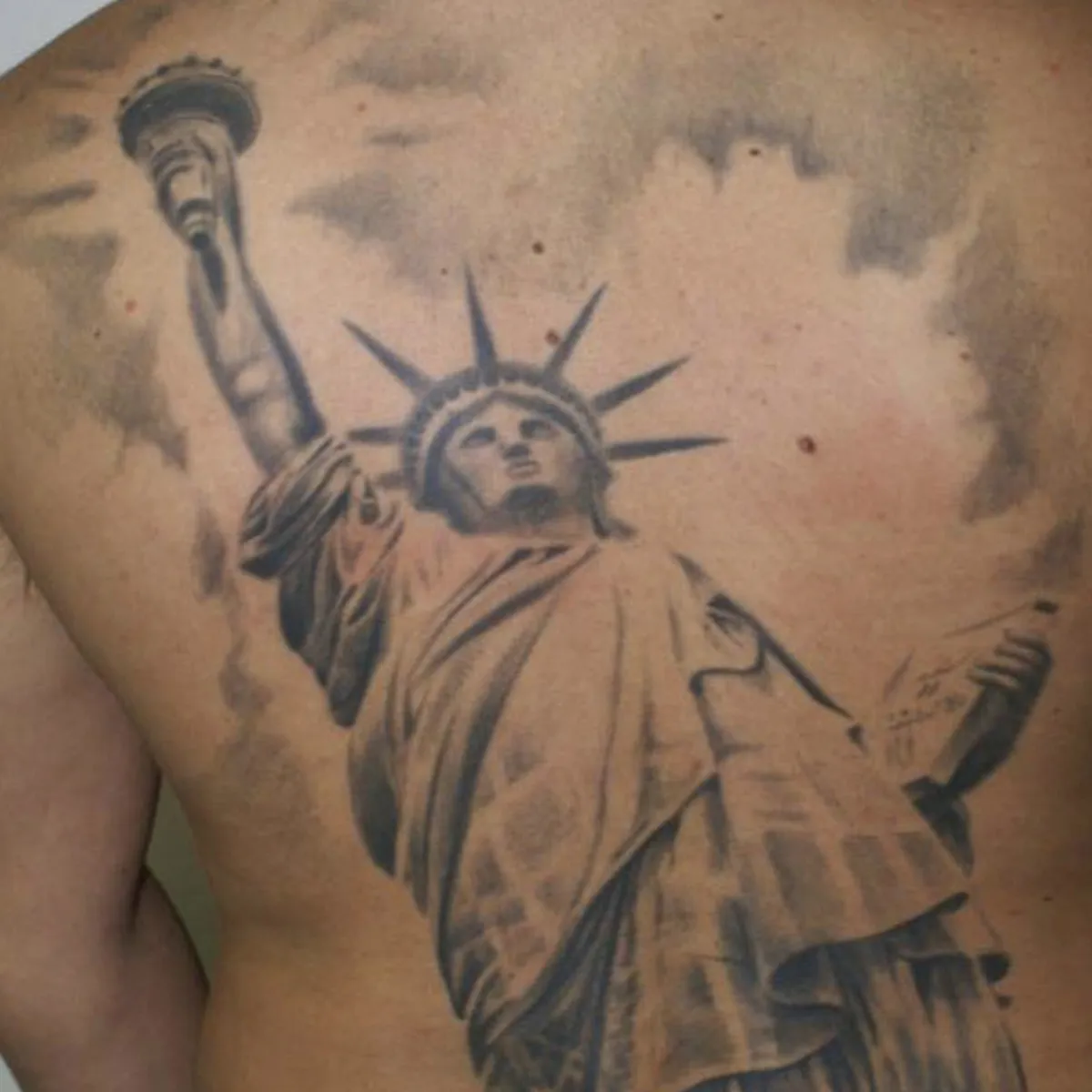 Tattoo of the Statue of Liberty with cloudy sky background on a person's back.