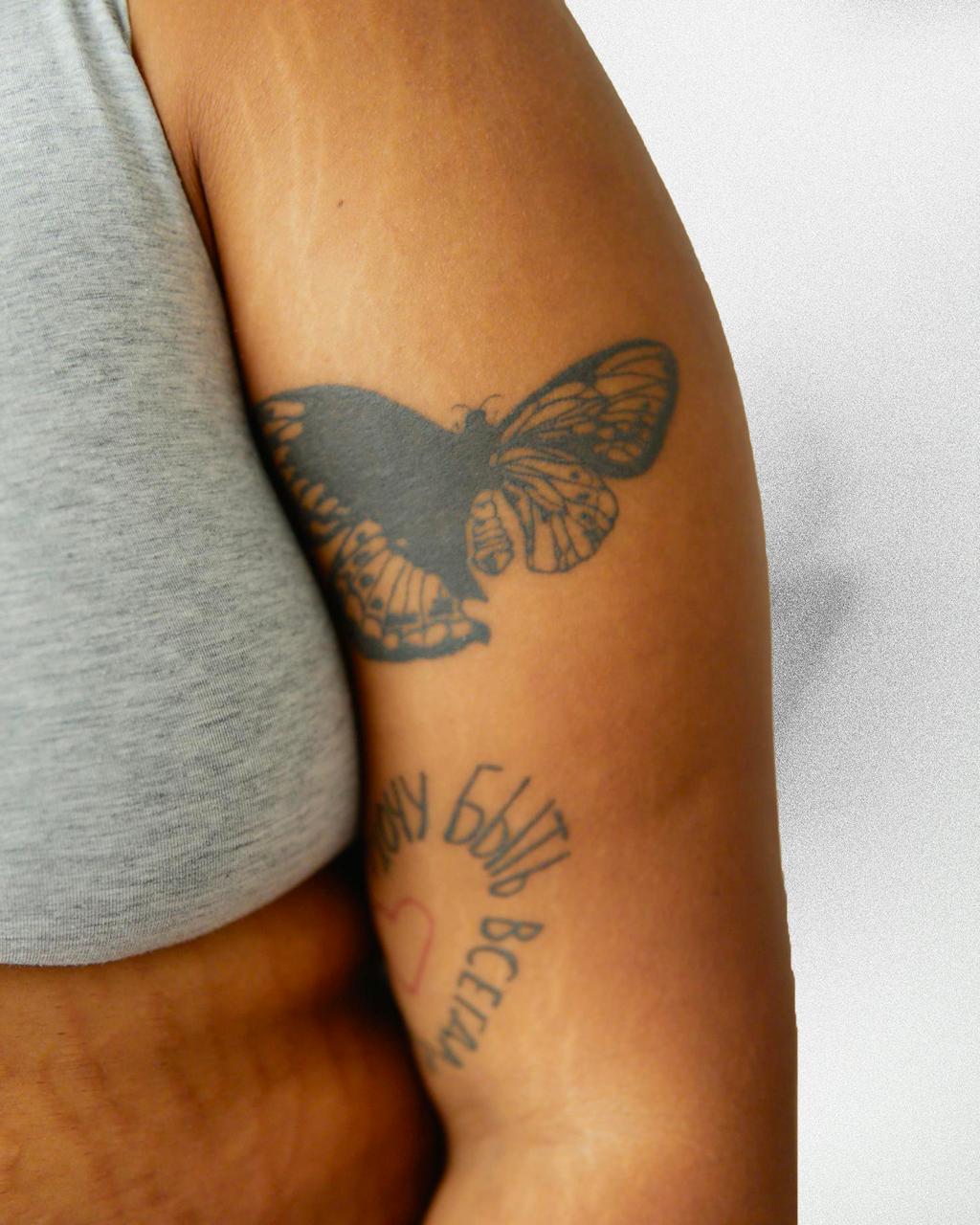 Close-up of a person's arm with a detailed black butterfly tattoo and partially visible cursive text tattoo below it.