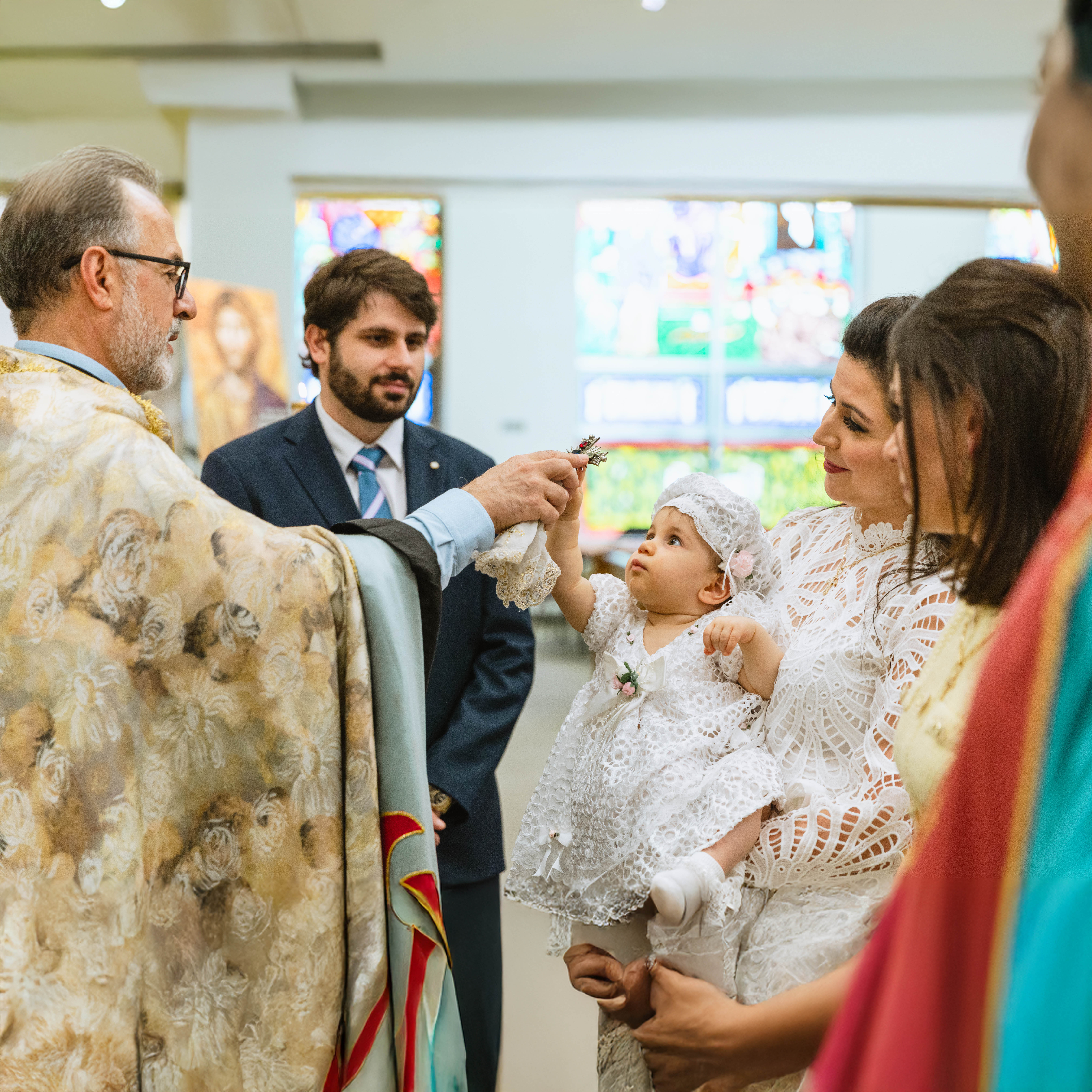 Priest holding a cross for a baby in white lace dress and bonnet held by a woman during a baptism ceremony in a church.